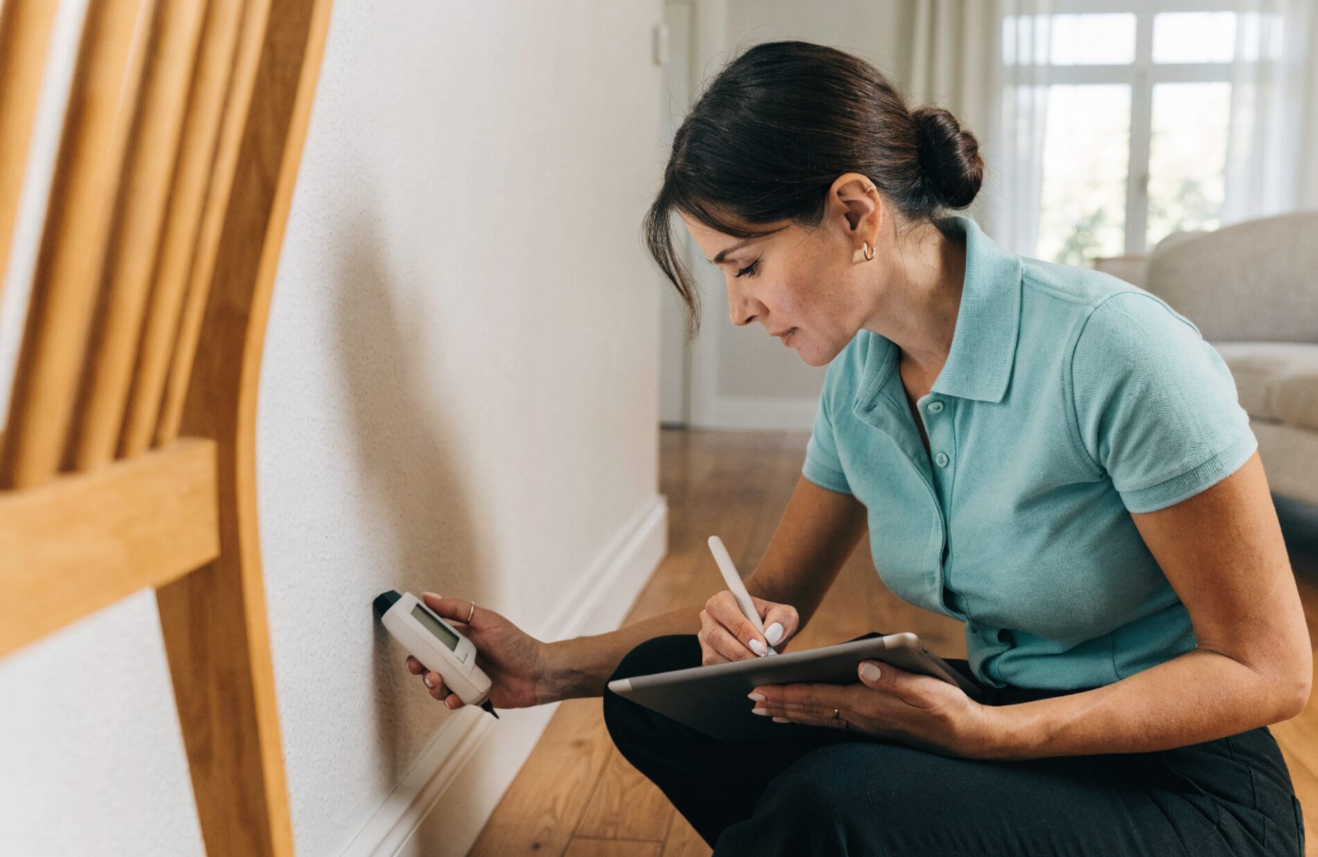 A mold inspector holding a moisture meter to a baseboard while recording findings on a tablet in a bright, clean home